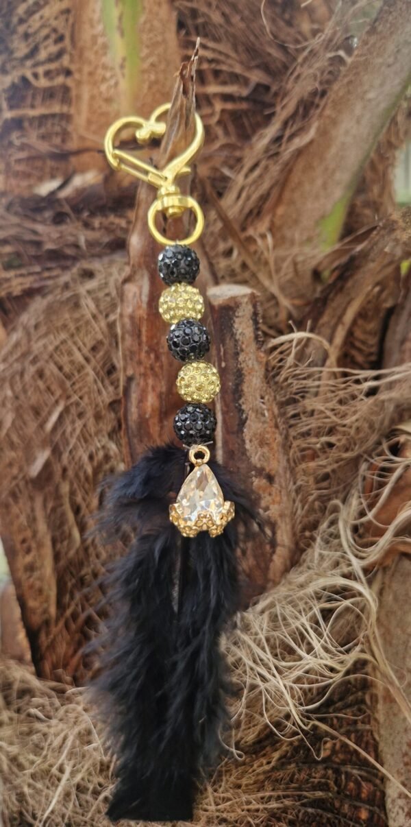 Een sierlijk halsterhanger ontwerp met een gouden hartvormige karabijnhaak, versierd met glinsterende zwarte en gouden kralen. Onderaan hangt een zwarte veer en een druppelvormige, heldere kristalhanger in een gouden zetting. De sleutelhanger is gefotografeerd tegen een natuurlijke achtergrond van bruin boomschors en vezels.
