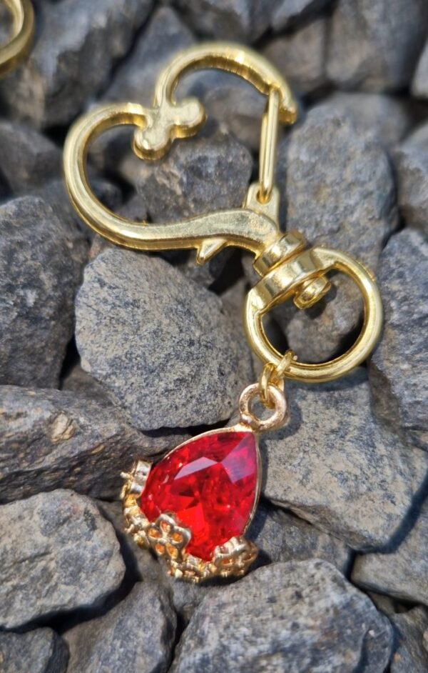 Een goudkleurige halsterhanger met een hartvormige clip, waaraan een rode, druppelvormige steen hangt in een sierlijke gouden setting. De halsterhanger ligt op een achtergrond van grijze stenen.