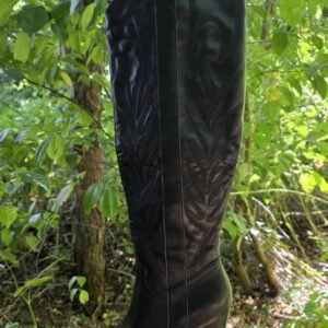 Zwarte westernlaars met geborduurd patroon en blokhak, gefotografeerd hangend tussen groene bladeren in een bosrijke omgeving.