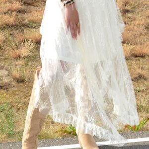 Vrouw met lang blond golvend haar draagt een lichtbruine cowboyhoed, een denim gilet en een witte, kanten gelaagde rok. Ze staat op een weg in een open duinlandschap met beige westernlaarzen en een ontspannen blik.