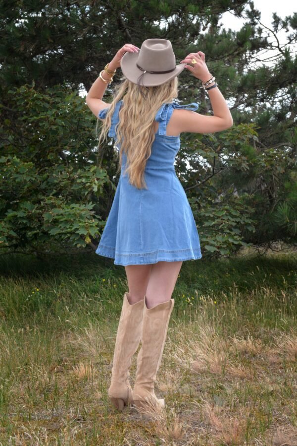 Model in lichtblauwe denimjurk met strikbandjes en beige suède cowboylaarzen in een open weidelandschap, met beige hoed en westernstijl accessoires.
