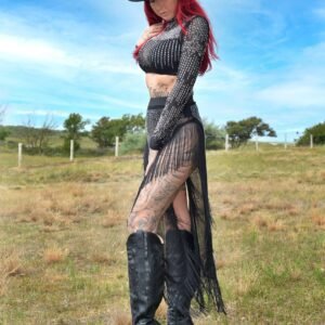 Model met rood haar in zwarte fringe-set met strass details, hoge zwarte laarzen en cowboyhoed in open grasland.