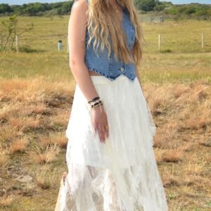 Vrouw met lang blond golvend haar draagt een lichtbruine cowboyhoed, een denim gilet en een witte, kanten gelaagde rok. Ze staat op een weg in een open duinlandschap met beige westernlaarzen en een ontspannen blik.