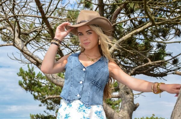 Blonde vrouw met groene ogen draagt een lichtbruine cowboyhoed, een mouwloos denim gilet en een witte rok met blauwe bloemenprint. Ze poseert voor een grillige boom in de duinen onder een heldere lucht.