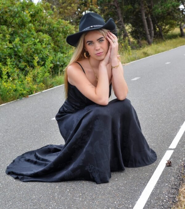 Vrouw in een zwarte lange jurk met subtiel bloemen en vlinderpatroon, zittend op een weg met een zwarte cowboyhoed en gouden sieraden, in een natuurlijke omgeving